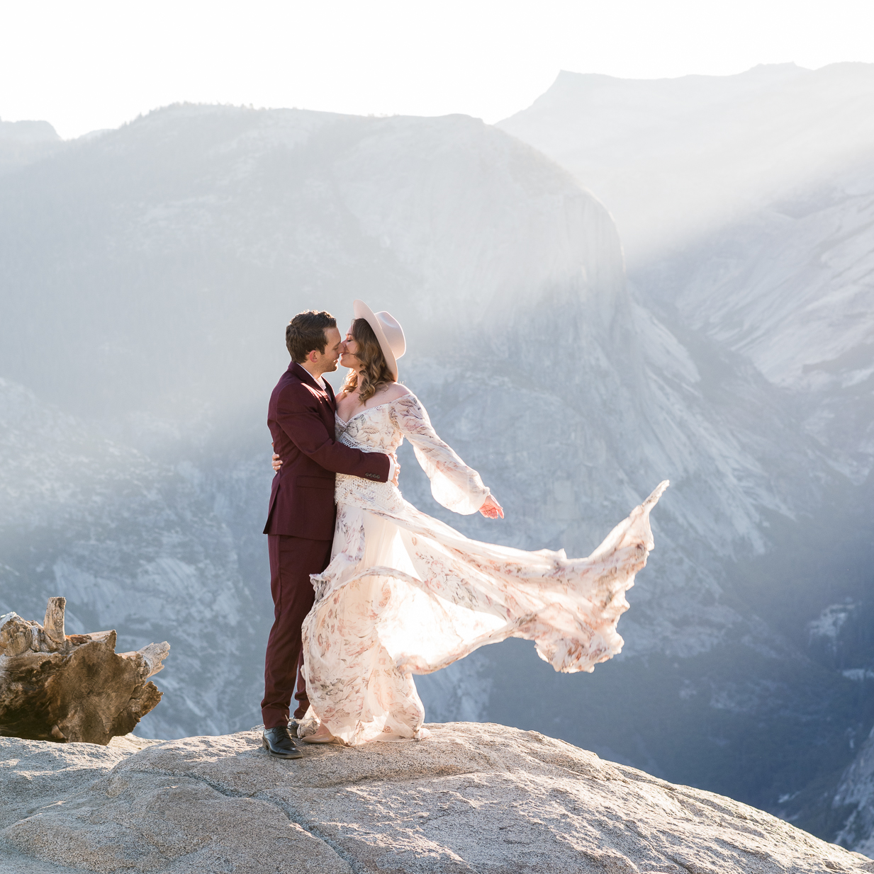 Elopement couple kissing on the edge of Glacier Point at sunrise in Yosemite National Park with beautiful light-rays and mountains as a backdrop.