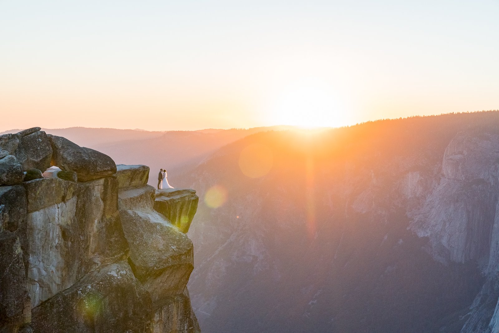 Epic Elopement Sunset photo with the bride and groom on the edge of a cliff at Taft Point, Yosemite National Park with the sun setting behind the valley rim creating a colorful sky.