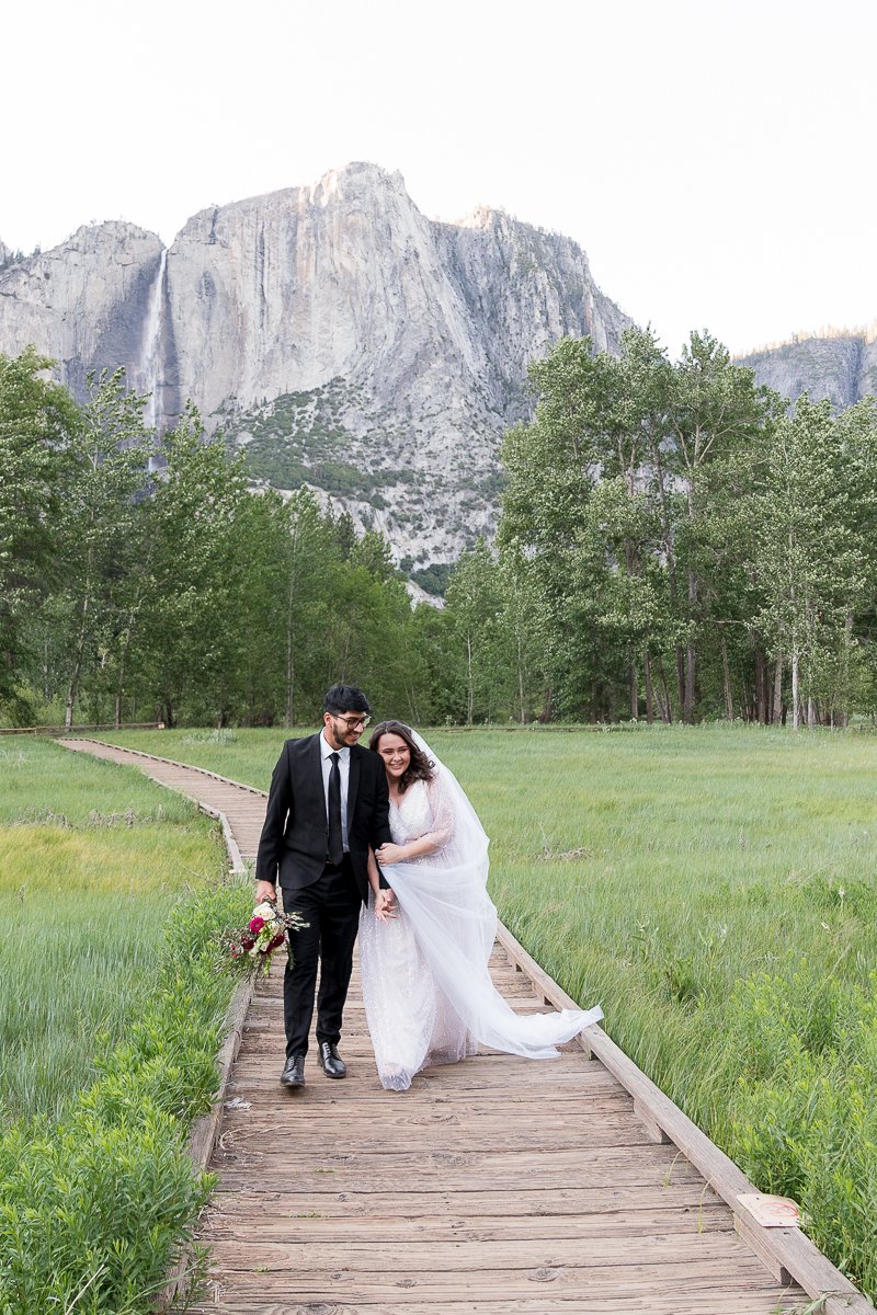 Elopement couple walking the wooden trail in the meadow of Yosemite Valley, Yosemite National Park with the waterfall in the background.
