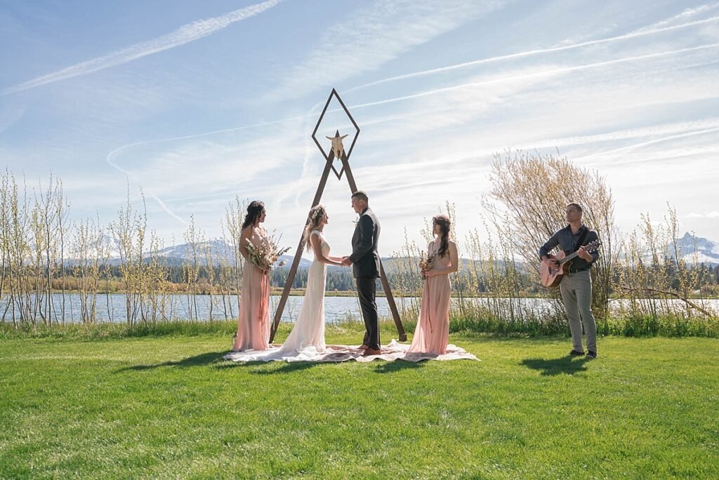 A bride and groom stand with two bridesmaids and a musician playing the guitar under an arch outdoors near a lake as they say their vows during their elopement.